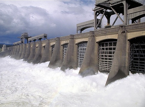 A large concrete dam stretches across white frothing water against a cloudy sky. 
