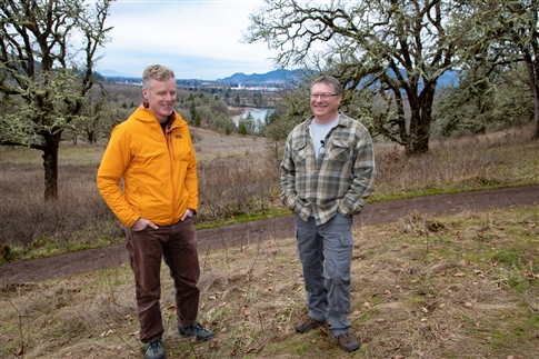 Two men stand outdoors with river in distance