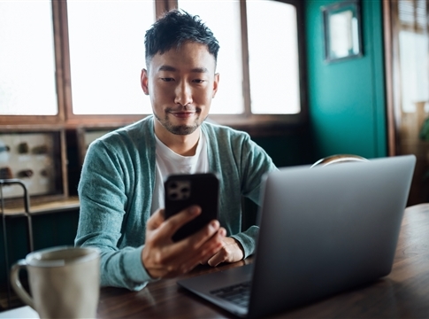 Confident young man looking at smartphone while working on laptop computer in home office. 